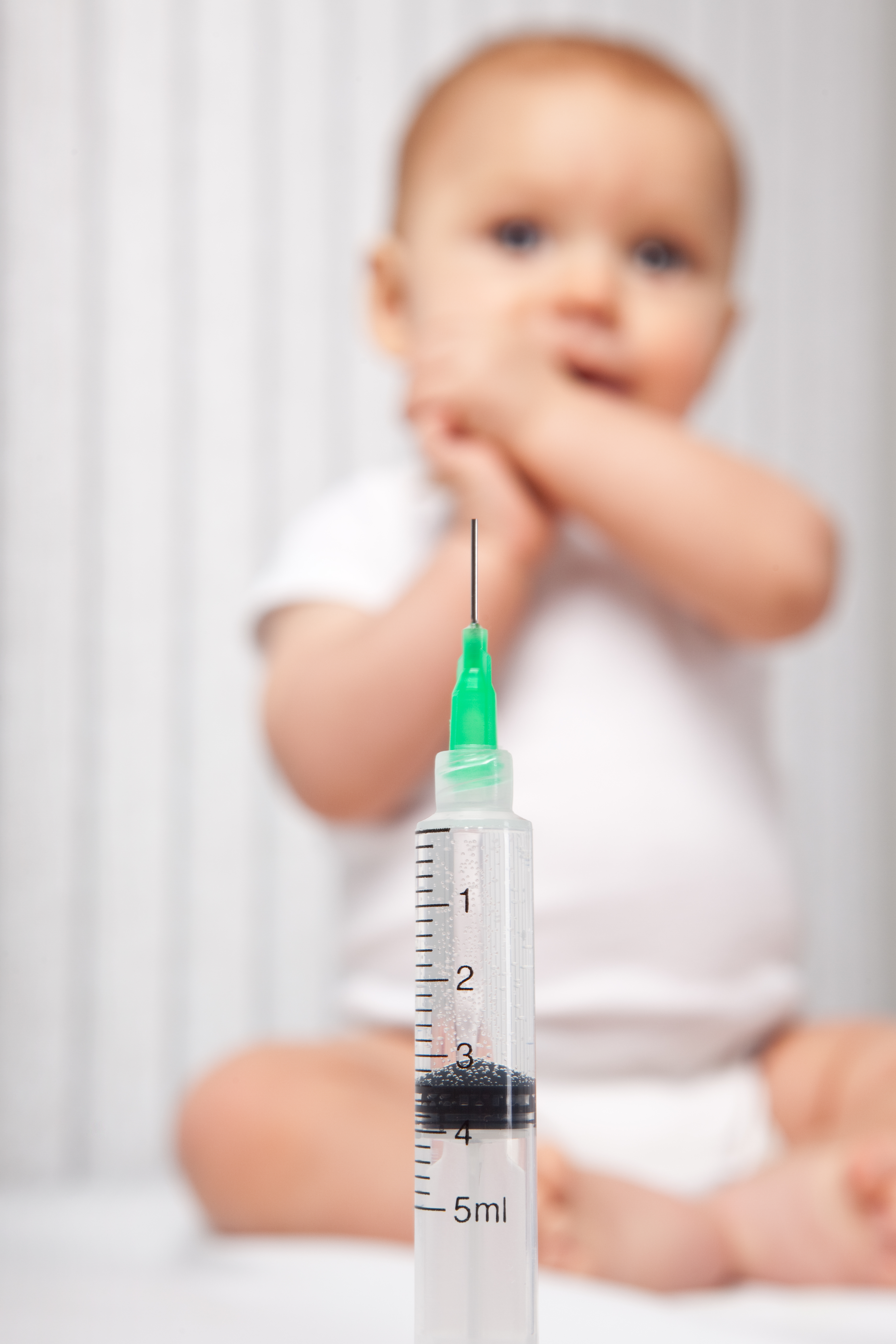 A syringe in focus with an 8 month old baby blurred in the background.